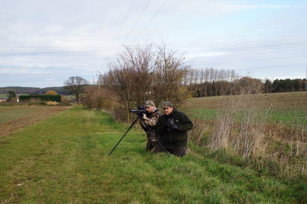 Zwei Jäger hocken auf einem Feld. Einer Blick durch das Zielrohr des Gewehr, welches auf einem Tripod aufgestellt ist.