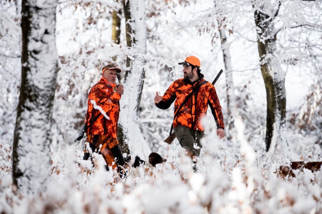 Zwei in orange gekleidete Jäger gehen durch den verschneiten Wald.