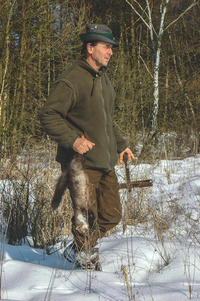 Jäger trägt getötetes Wiesel und Falle durch verschneiten Wald.
