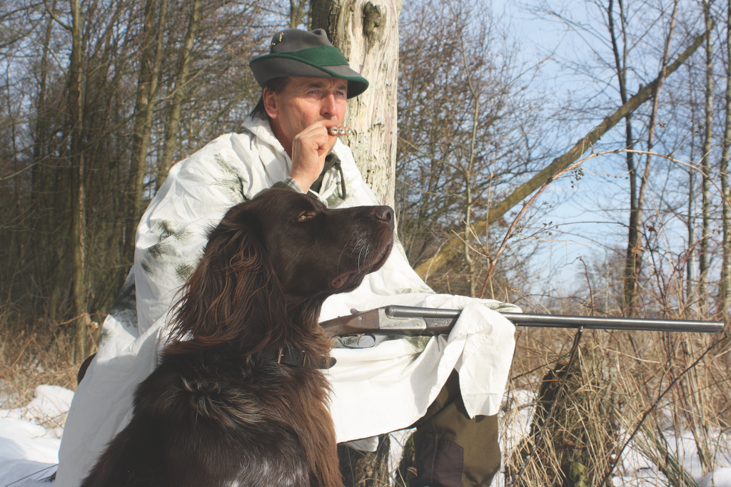 Jäger bläst in Fuchsflöte. Jagdhund sitzt neben ihn.